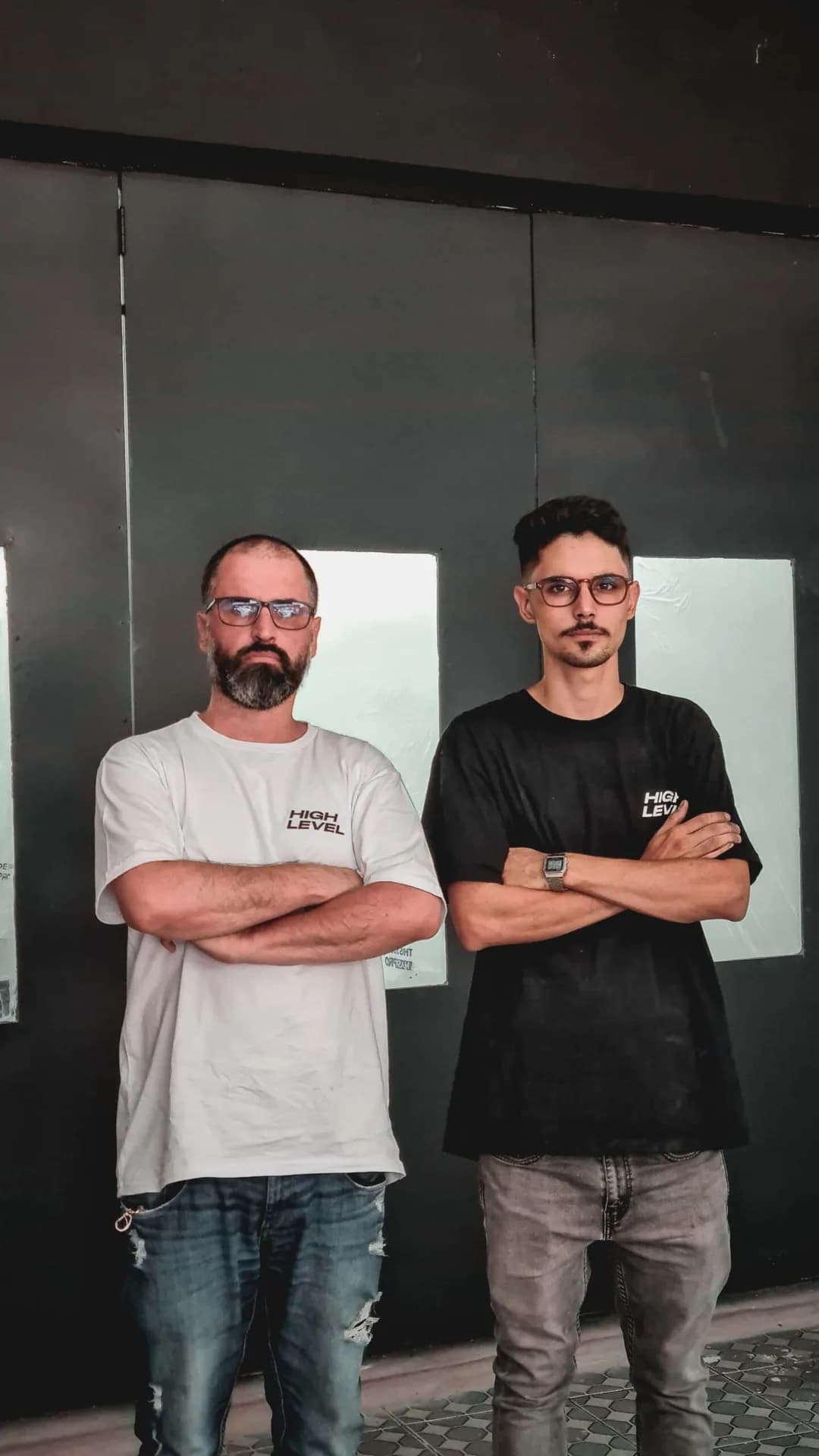 Dois integrantes da equipe da High Level de braços cruzados, com camisetas da oficina